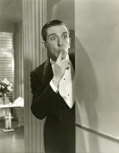 Man peeking out of a corner with his finger over his mouth saying "shhhh"