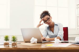 Deadline stress concept - sad african-american business woman sitting at desktop in office and holding hand on head. Hard working day.