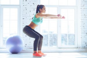 Deep squat. Side view of young beautiful woman in sportswear doing squat and holding dumbbells while standing in front of window at gym