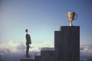 Business man standing at the bottom of steps that lead to a golden trophy