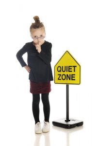 An adorable little girl school teacher or librarian, gesturing to silence the viewer. She stands in front of a "Quiet Zone" sign. On a white background.