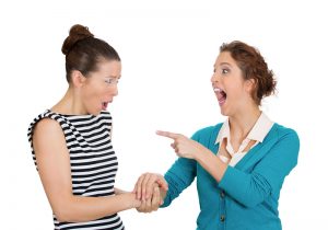 Two women with shocked expressions, pointing at another while conversing