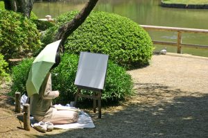 Person sitting on a blanket in the park with a parasol and an easel