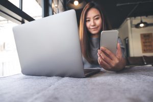 Closeup image of a beautiful Asian woman holding and looking at mobile phone while using laptop in cafe