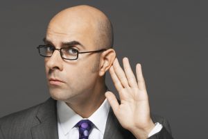 Closeup of a bald businessman with hand behind ear listening closely against gray background